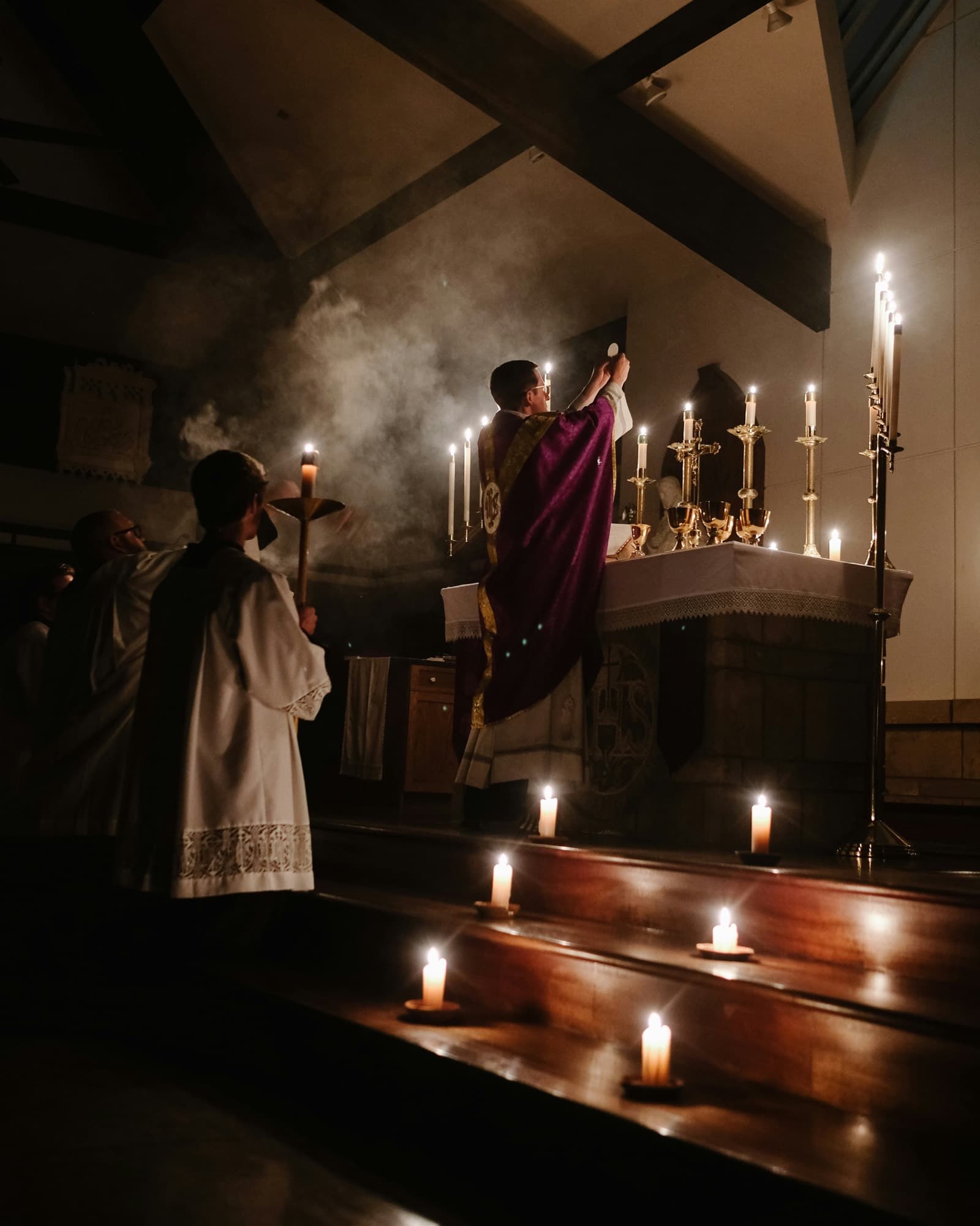 A priest in a purple chasuble elevating the host at a candlelit altar, with servers in white beside him.