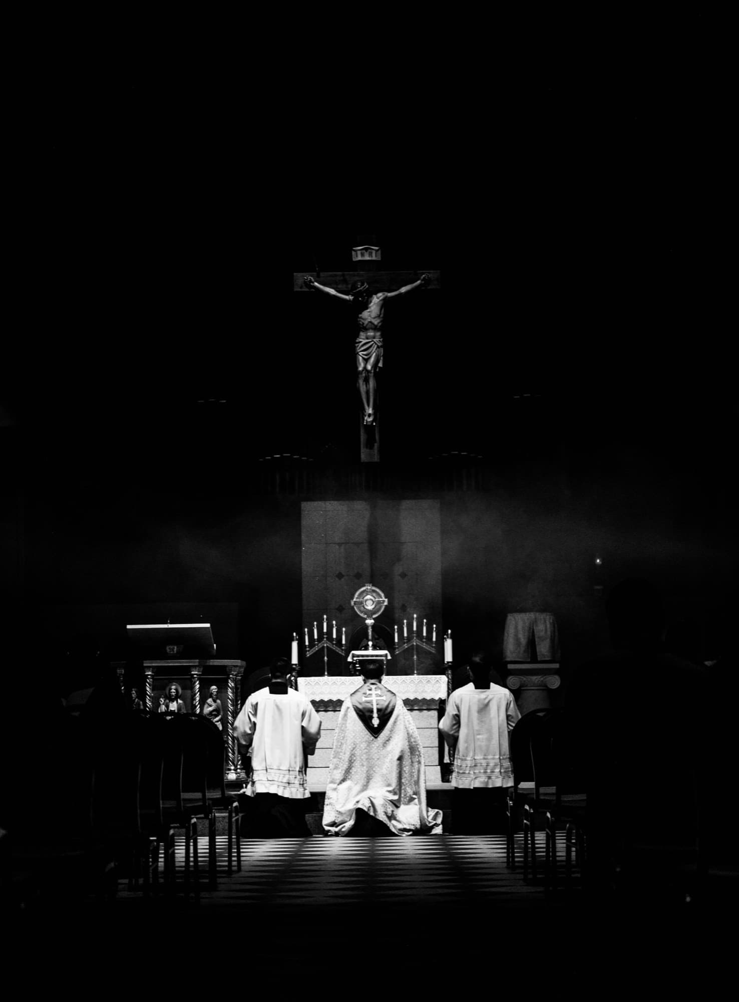 Three clergy kneeling before an altar beneath a crucifix in a dark church.