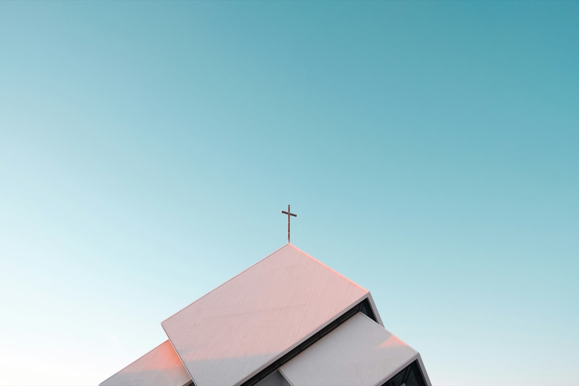 A cross on the peak of a pale church roof against a clear blue sky.