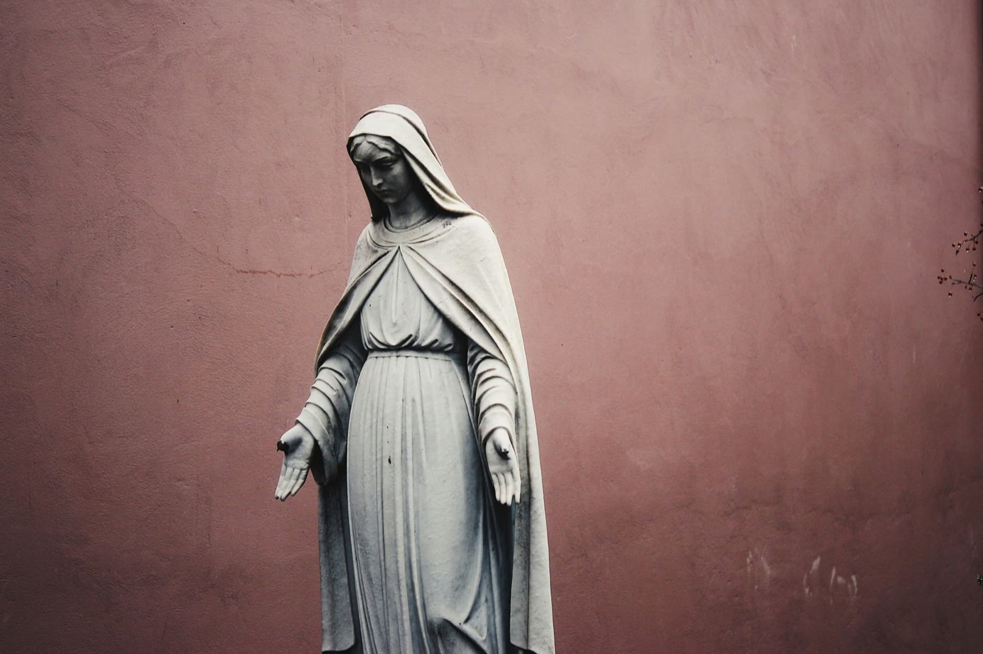 A stone statue of Mary with open hands against a rose-colored wall.