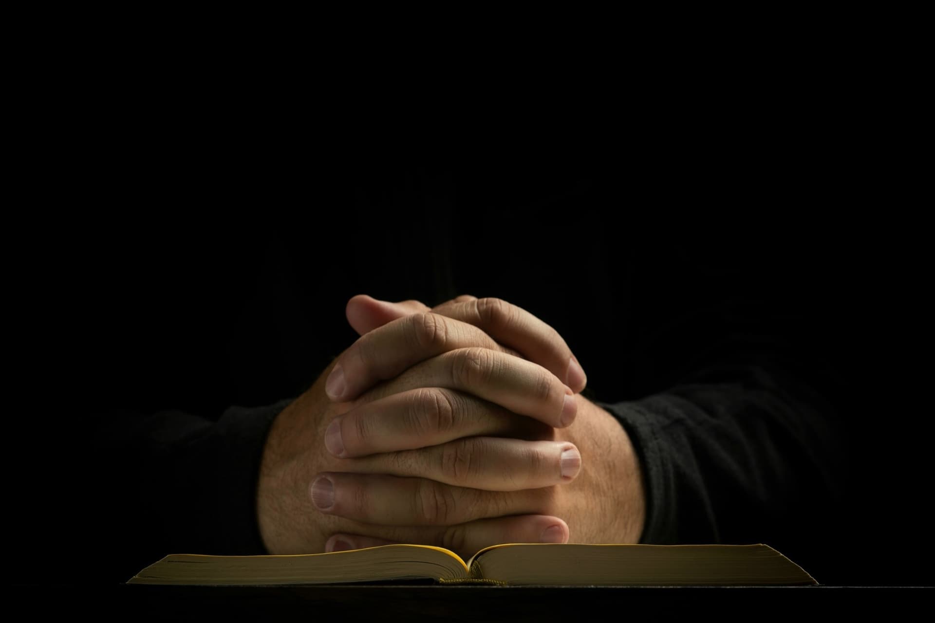 Folded hands above an open book in a dark room.