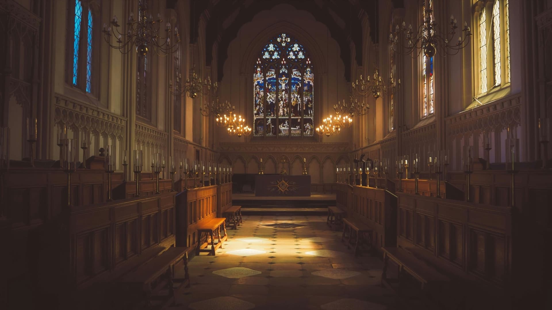 A candlelit chapel with wooden stalls, chandeliers, and a stained-glass window above the altar.