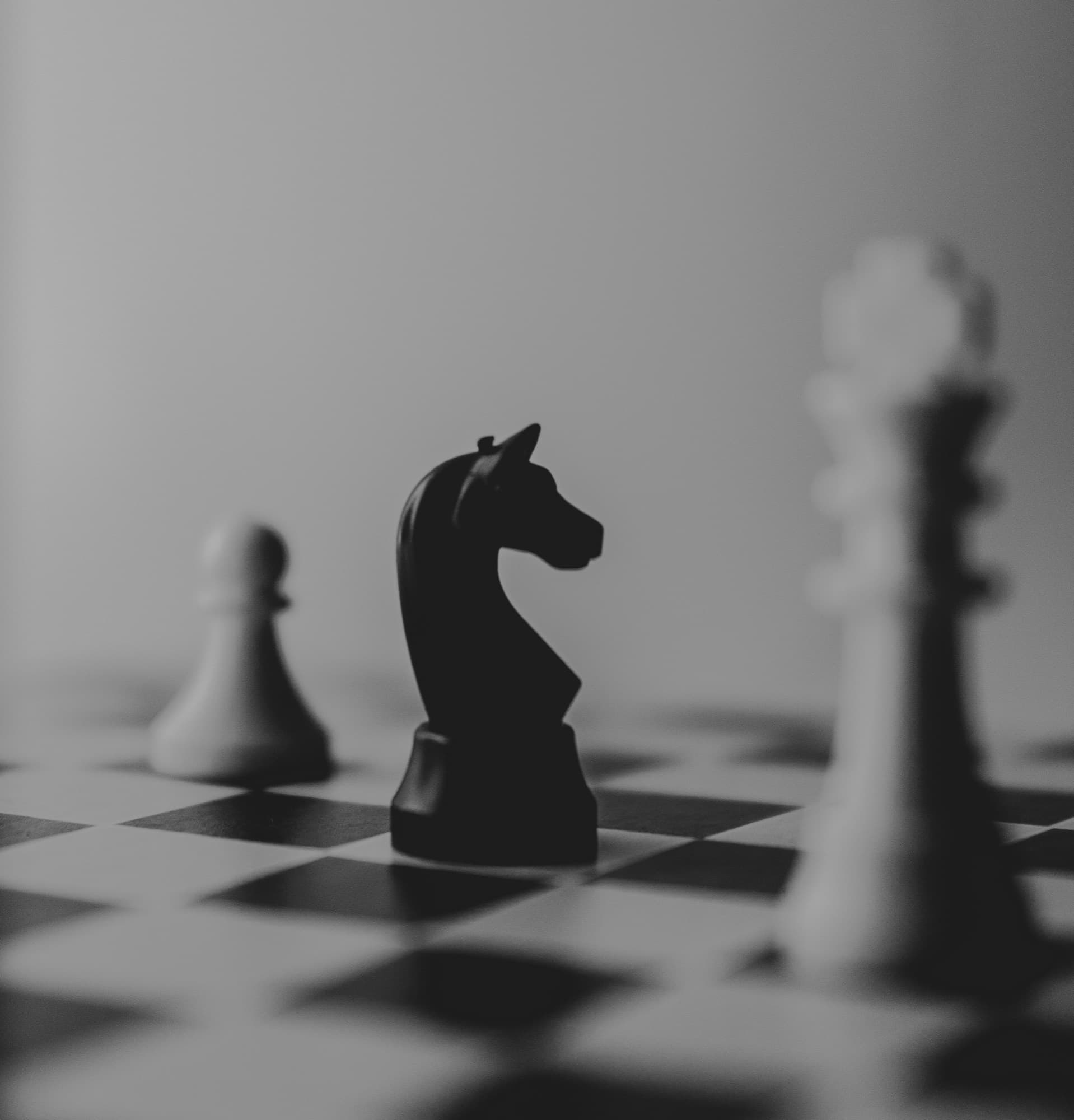 A black knight chess piece in focus on a board, with a pawn and a king blurred behind it.